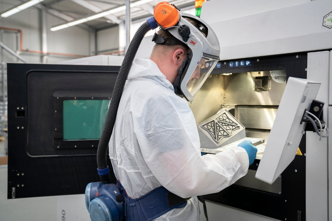 Ein Mann in Schutzausrüstung entnimmt ein Bauteil aus dem 3D-Drucker